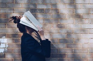Woman holding an open book over her face. Is she upset by all the unnecessary backstory in the novel?